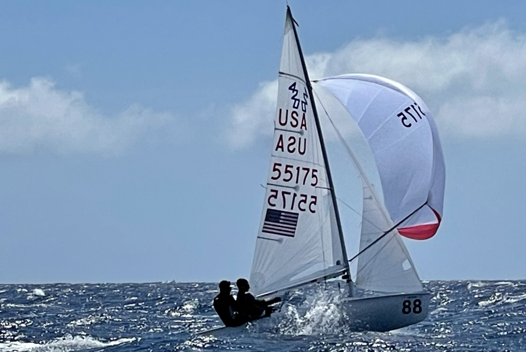 Two people are sailing a small boat with sail number 55175 on the ocean. The sail displays "USA" and an American flag. The boat is leaning, and the sky is partly cloudy.
