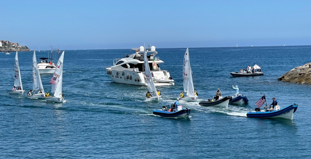 Several small sailboats and motorboats with people onboard move near a large white yacht on a calm blue sea, with a rocky outcrop and another boat in the background under a clear sky.