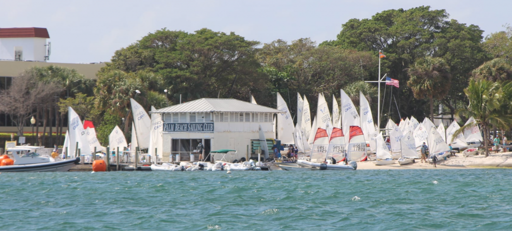 A marina scene shows a sailing club building surrounded by numerous small sailboats with white sails along the shore, trees in the background, and turquoise water in the foreground.