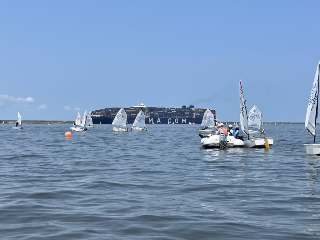 Small sailboats with people on board float in the foreground on calm water, while a large cargo ship labeled “CMA CGM” crosses the background under a clear blue sky.