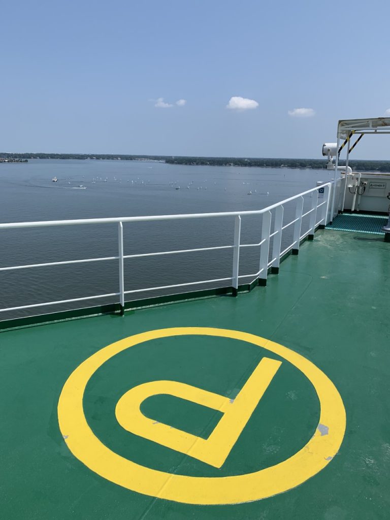 View from a ship’s deck with a yellow helicopter landing circle on green floor, white railings, calm water, and a distant shoreline under a clear, blue sky with a few clouds.