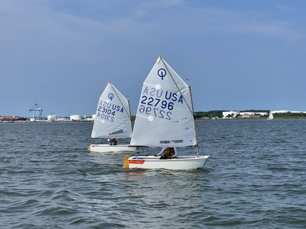 Two small sailboats with white sails, each marked "USA" and numbers, are being sailed by individuals on a calm body of water. There is a port and industrial structures visible in the distant background.