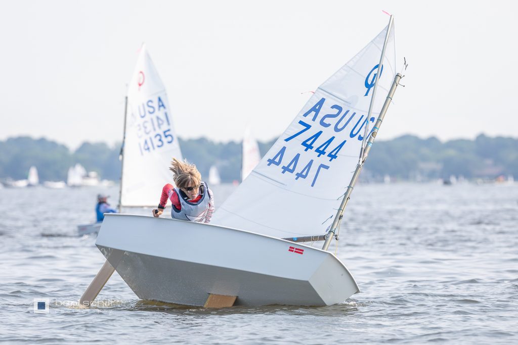 A young sailor leans out of a small white sailboat marked “USA 7444” on a calm lake, surrounded by other sailboats in the background under a hazy sky.
