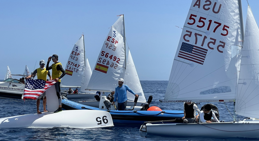 Sailors on small boats gather on the water, with some standing on an overturned boat holding an American flag. Other boats display USA and Spain sail numbers. The scene is sunny and calm.
