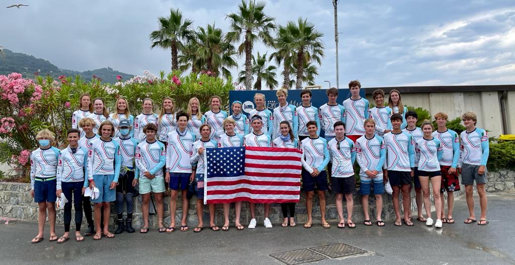 A large group of young people in matching white, blue, and red tops stands outdoors holding an American flag. Palm trees, plants, and a cloudy sky are in the background. They appear to be part of a team or group event.