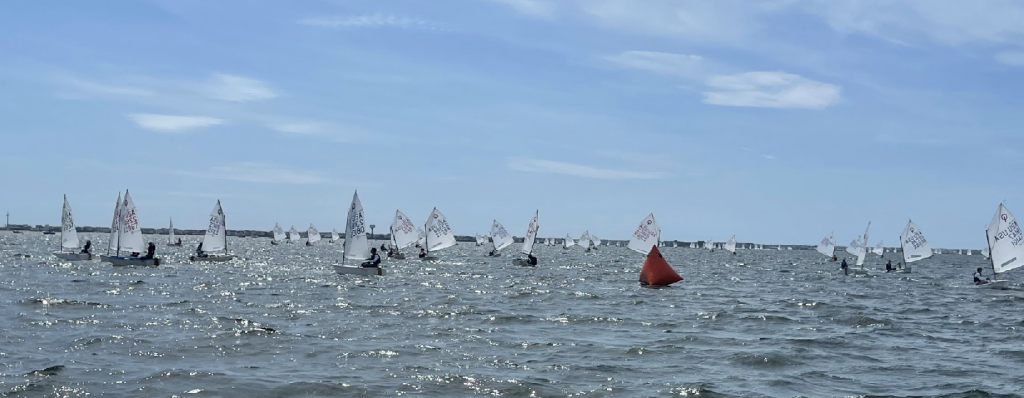 A group of small sailboats with white sails race on a sunny, windy day. The boats are spread out across choppy water, with a red buoy visible near the center. The sky is mostly clear with some clouds.