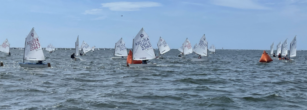 A group of small sailboats with white sails compete in a regatta on a choppy body of water, passing orange buoys under a partly cloudy sky.