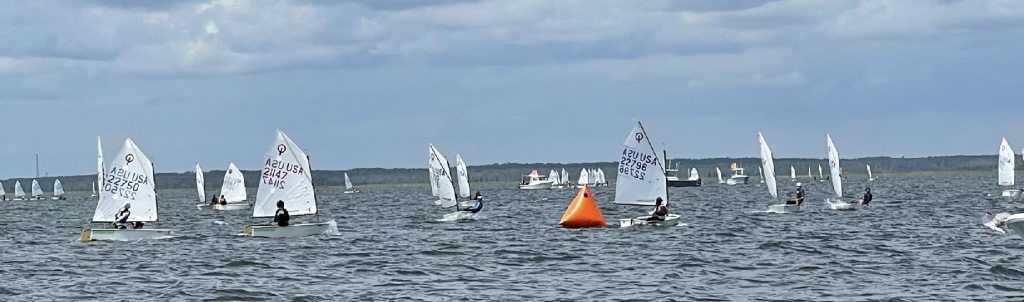 A group of small sailboats with white sails participates in a regatta on a choppy lake, moving around an orange buoy under a cloudy sky.