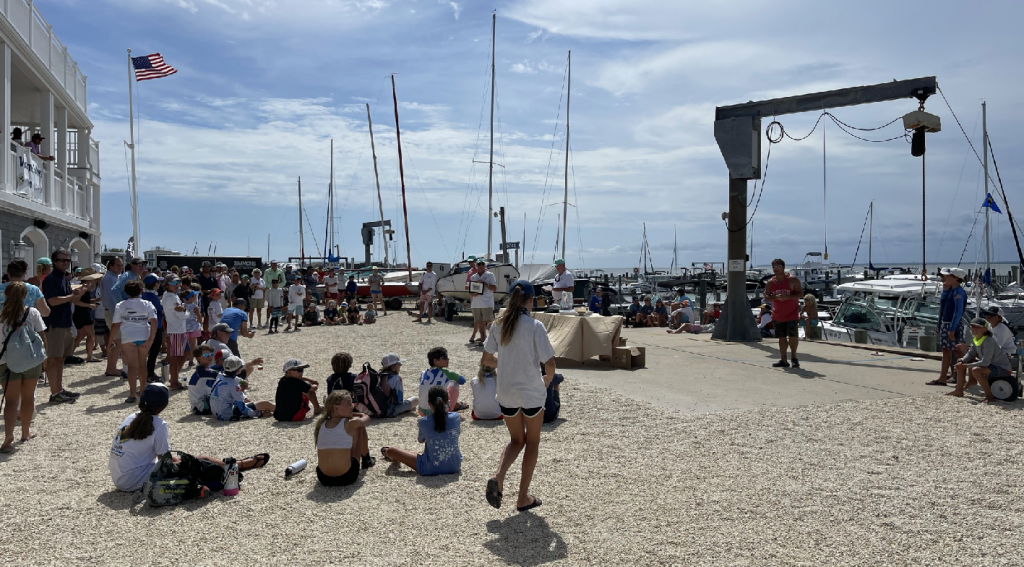 A crowd of people, many in casual summer clothes, gather near sailboats and docks at a marina on a sunny day. Some sit on gravel while others stand, with an American flag waving on a building to the left.