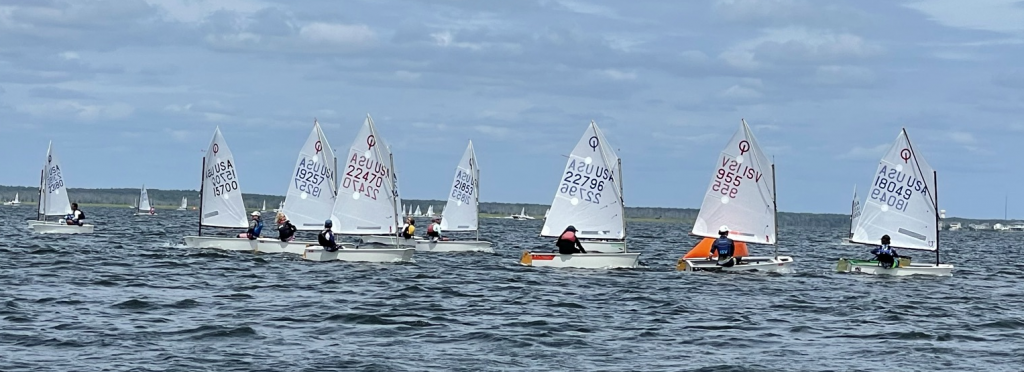 A group of young sailors in small sailboats races across a wavy body of water under a partly cloudy sky, with land visible in the background.