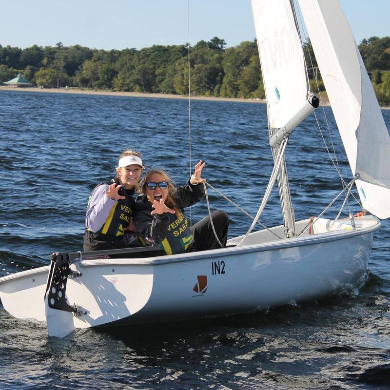 Two people wearing life jackets and sunglasses smile and gesture with peace signs while sitting in a small sailboat on a lake, with trees and shoreline visible in the background.