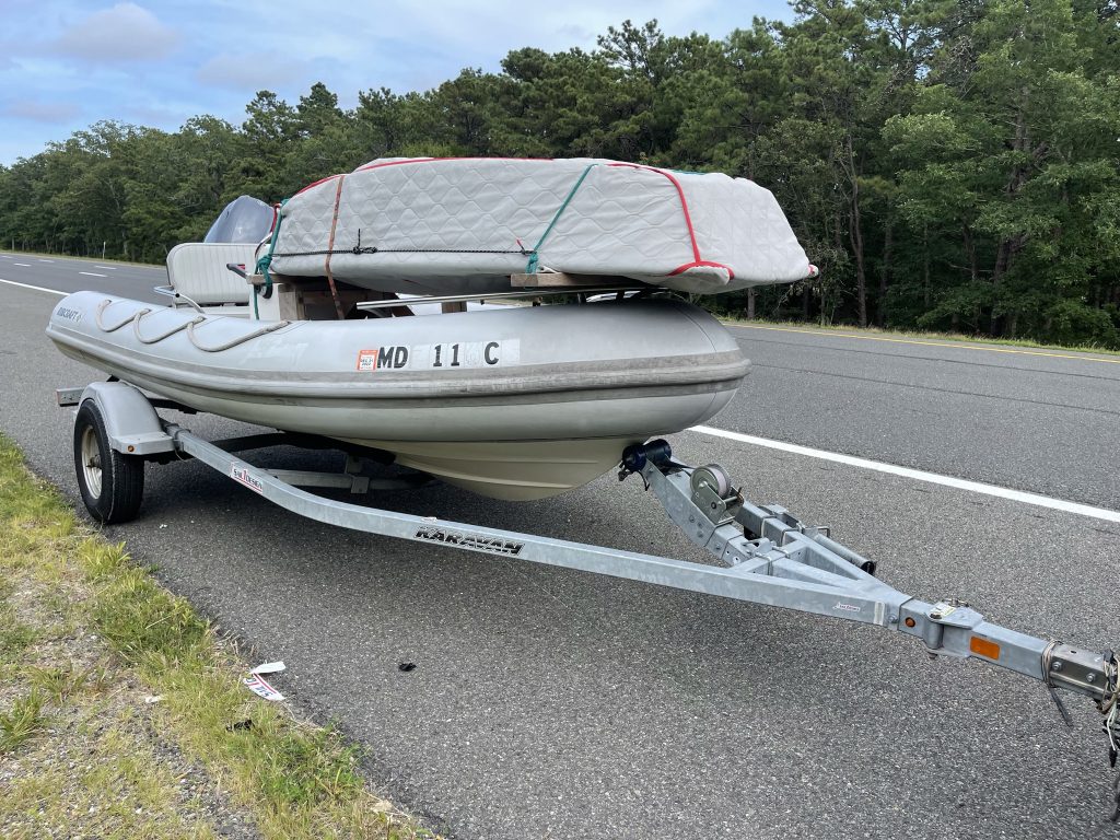 A grey inflatable boat on a trailer is parked on the side of a highway. A mattress wrapped in plastic and tied with red and green straps is secured on top of the boat. Trees line the road in the background.