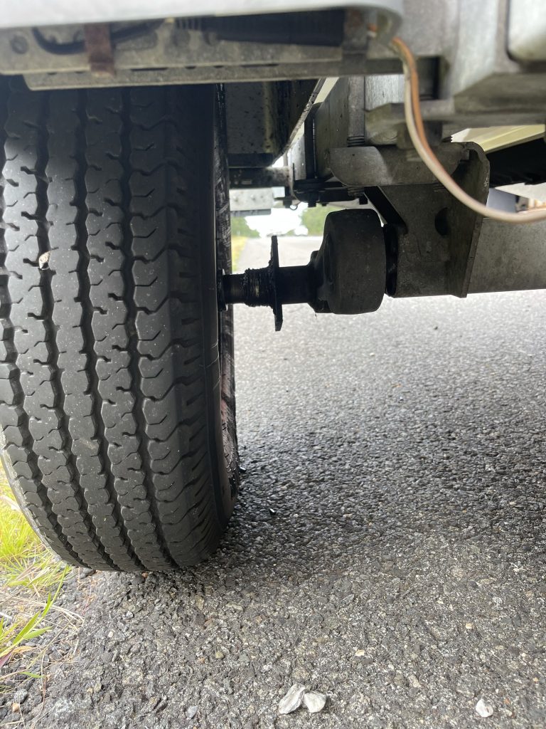 A close-up view of a vehicle’s rear tire and axle on a paved road, showing the wheel visibly separated from the damaged axle, indicating a possible mechanical failure.