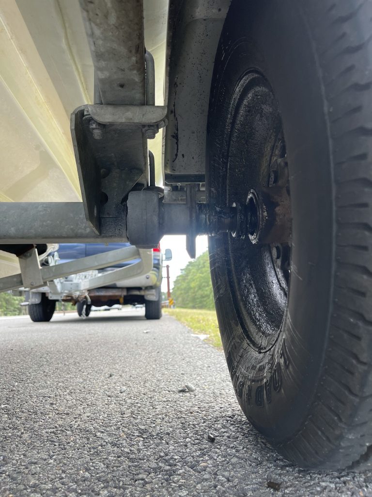 Close-up view of a black trailer wheel and axle on a road, with a blurred vehicle and boat trailer visible in the background. The ground is asphalt, and there is greenery along the side of the road.