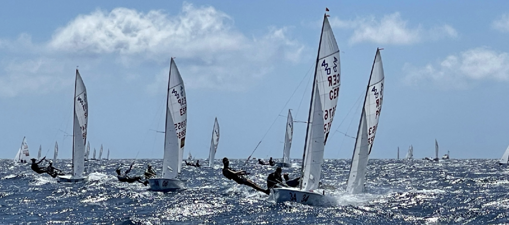 Sailboats with raised sails race across a choppy blue sea under a partly cloudy sky. Several sailors lean out over the water, balancing their boats as they compete.