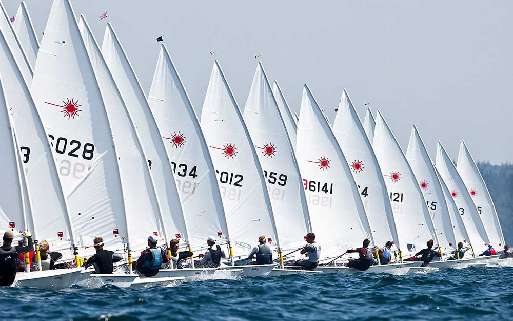 A group of sailors in individual small sailboats with white sails, lined up close together on blue water, ready for a race. The sails display numbers and a red sunburst emblem. It is a sunny day.