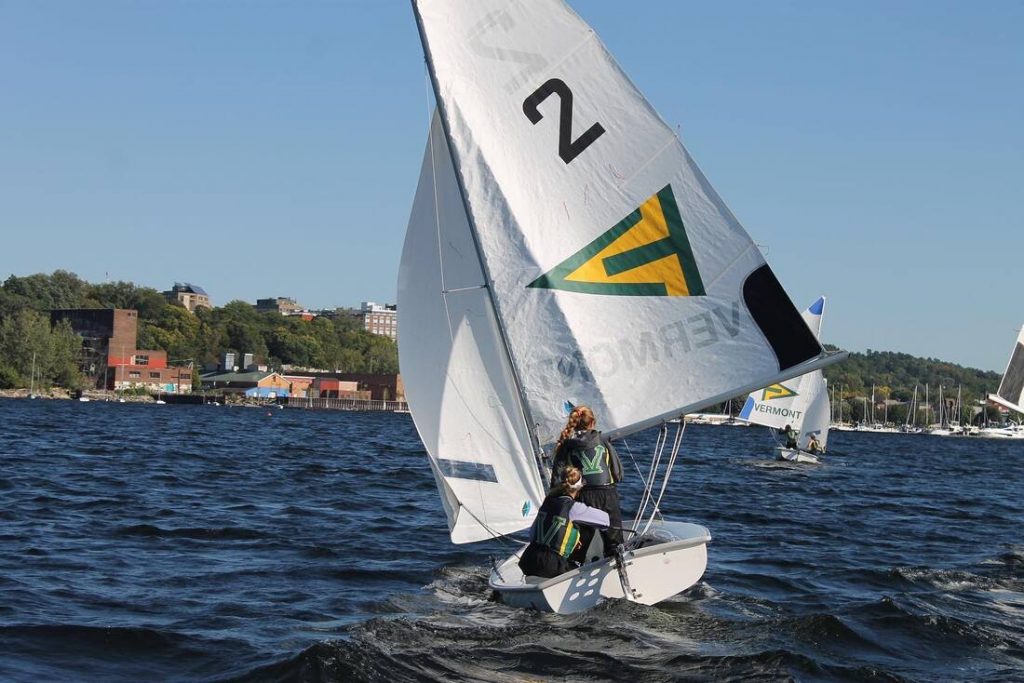 Two people sail a small white boat with a '2' on its sail across a blue lake. Other sailboats and a shoreline with buildings and trees are visible in the background under a clear sky.