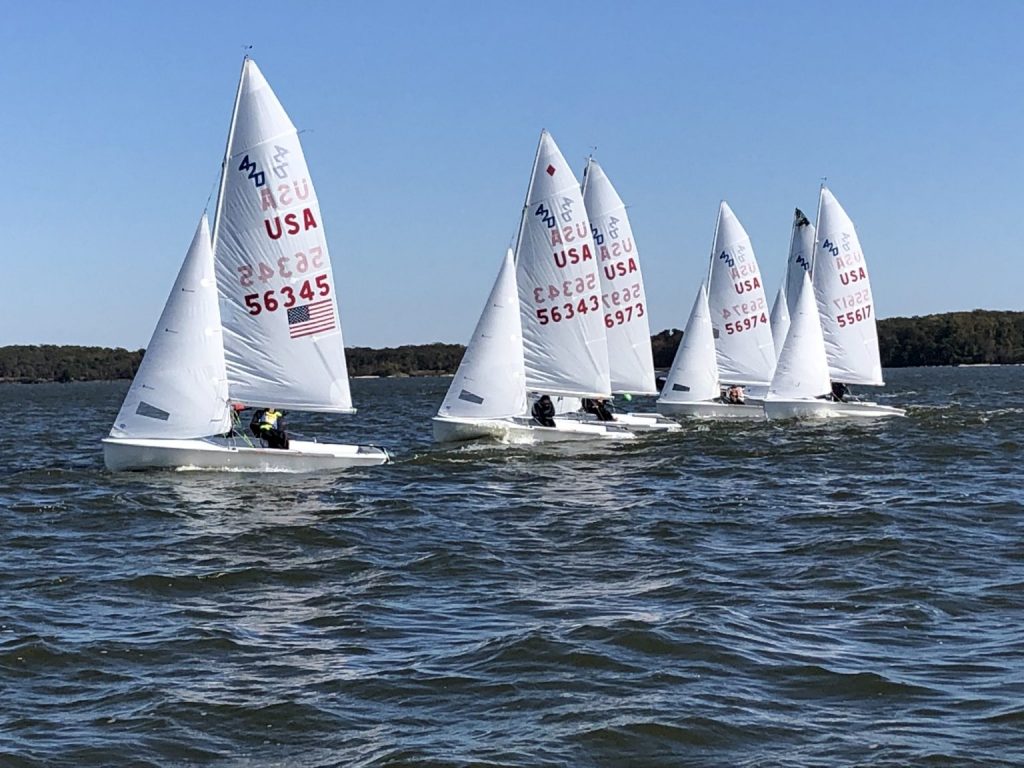 Five small sailboats with white sails, marked with USA and numbers, race closely together on a lake under a clear blue sky, with tree-lined shore in the background.