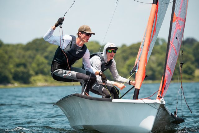 Two people wearing sunglasses and sailing gear lean out of a small sailboat, holding ropes, as they navigate on a lake with a forested shoreline in the background.