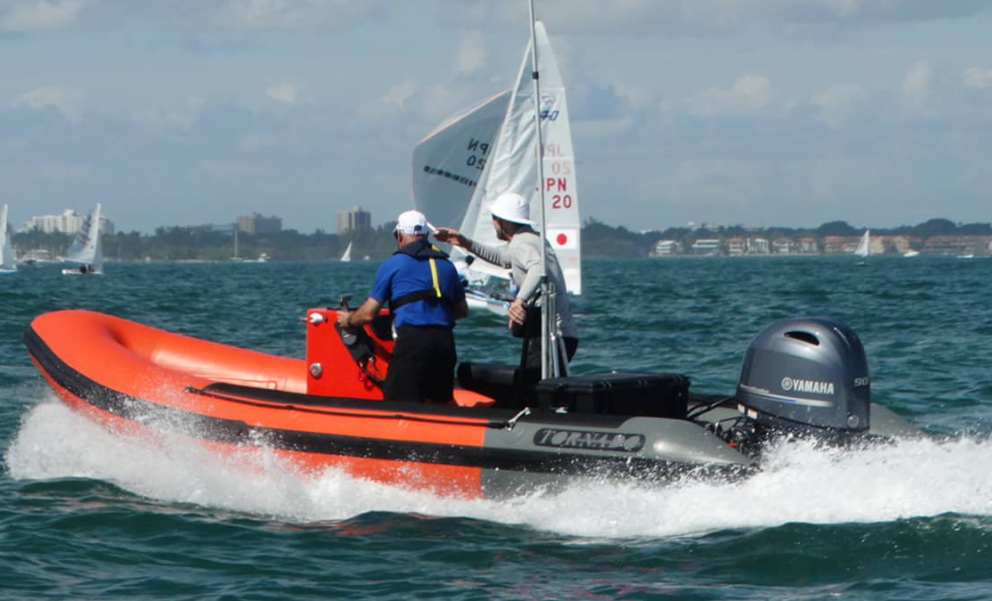 An orange and gray Yamaha motorboat speeds through the water, carrying two people in hats and life jackets. One person points ahead, while sailboats and a shoreline with buildings are visible in the background.
