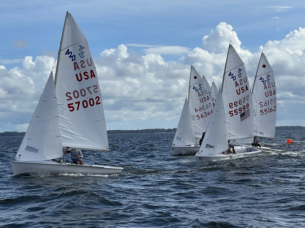Four sailboats with white sails and 'USA' markings race on choppy water under a partly cloudy sky. The boats are close together, tilted by the wind, with a distant shoreline visible in the background.