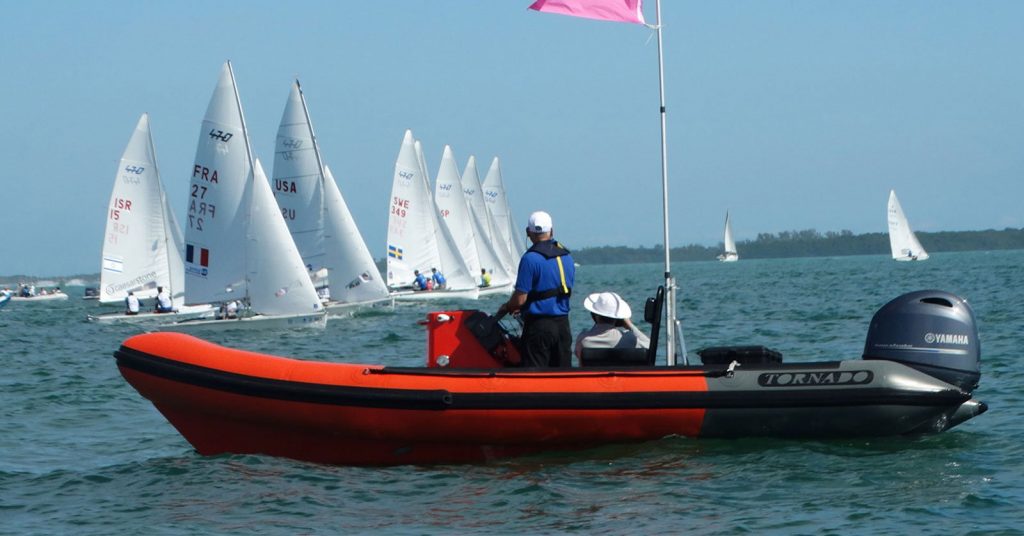 A person in a blue shirt stands on an orange and black inflatable motorboat with a pink flag, observing a sailing race with several white sailboats on the water in the background.