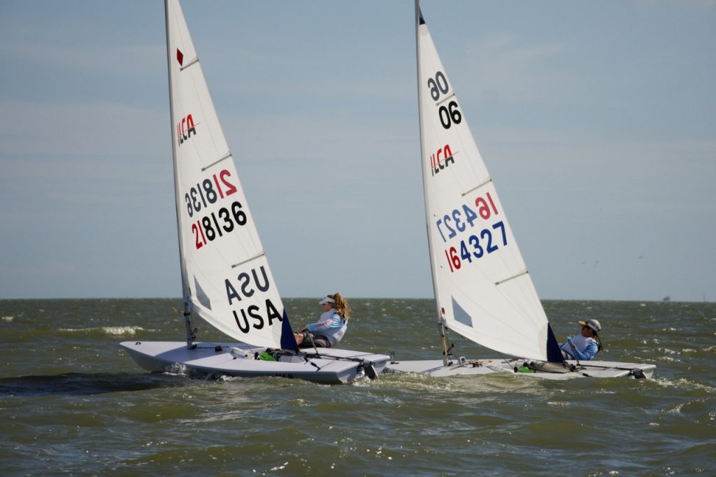 Two people sail small racing sailboats, marked 'USA 218136' and 'USA 64327,' side by side on wavy water under a blue sky. Both sailors wear blue shirts and hats, leaning back to balance their boats.