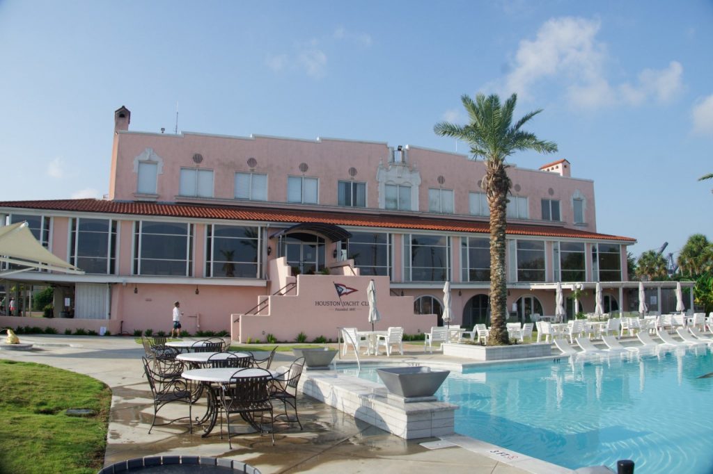 A large pink building with arched windows stands behind an outdoor swimming pool, surrounded by white lounge chairs, tables with chairs, palm trees, and a bright blue sky.