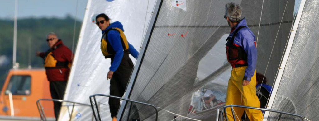 Three people stand on the decks of sailboats, wearing life jackets and facing different directions. The sails are raised, and a small orange boat is visible in the background on the water.