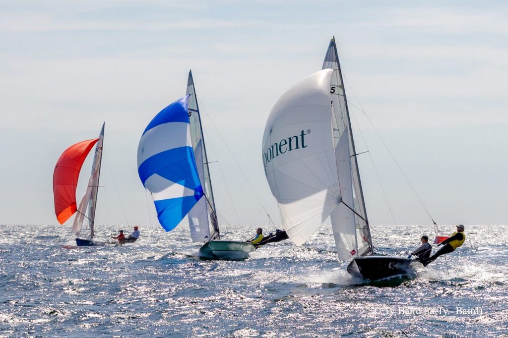 Four sailboats with colorful spinnakers—red, blue, and white—race across a sparkling sea under a bright sky, with crews leaning out to balance the boats.