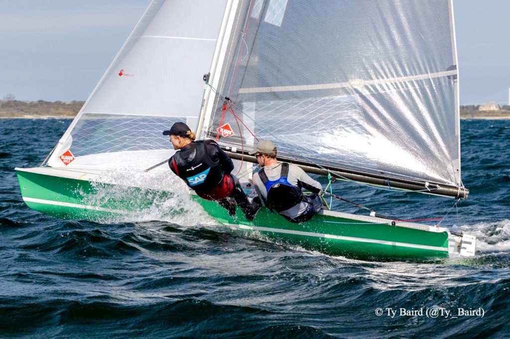 Two sailors maneuver a green racing sailboat on choppy water, leaning out to balance as the sail catches wind. Both wear life jackets and caps; one sailor is branded with 'SAP.' The sky is clear and land is visible in the distance.