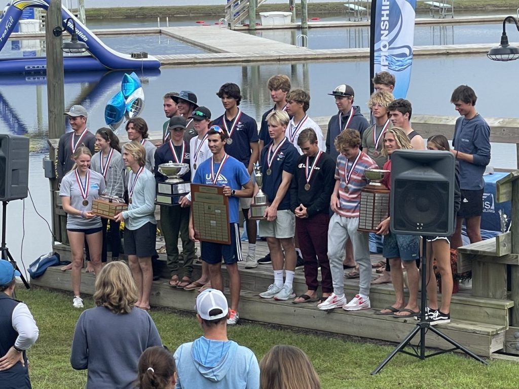 A group of young people with medals stand on a dock holding trophies and plaques, posing for a photo at an outdoor awards ceremony near the water, while spectators watch.