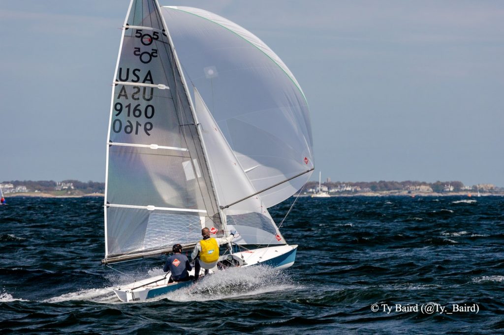 Two sailors wearing life jackets navigate a small sailboat with a large white sail, marked "USA 9160," through choppy blue water near a distant shoreline under a partly cloudy sky.