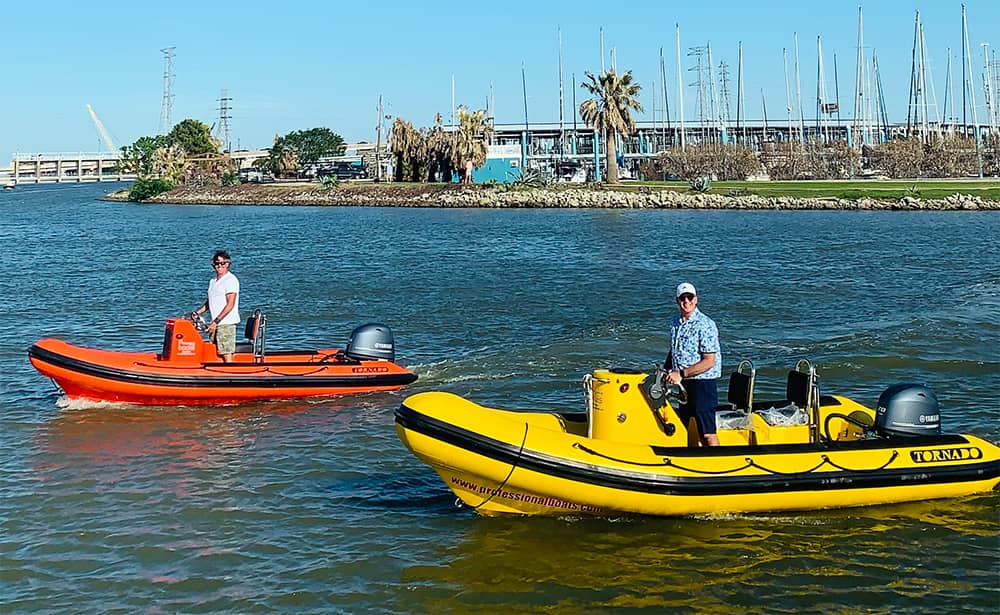 Two people each steering small inflatable motorboats—one orange, one yellow—on a body of water near a shoreline with palm trees, grass, and sailboat masts in the background under a clear blue sky.