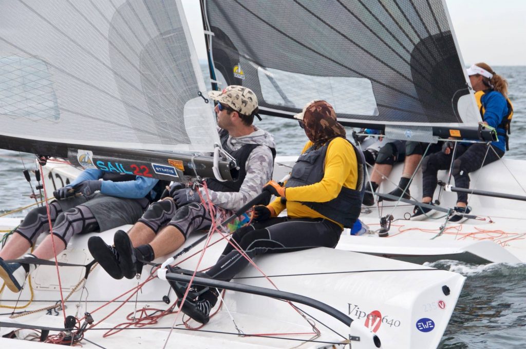 Four people are sailing two small racing sailboats close together, wearing life vests and outdoor gear, while adjusting ropes and leaning out over the water during a regatta.