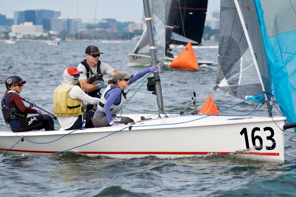 Four people sailing a small white boat numbered 163 in a race, adjusting ropes and sails, with city buildings and other boats visible in the background on the water.
