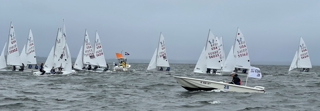 Small sailboats with numbered sails line up near a yellow flag boat in choppy water, while a motorboat with two people and the number 28 leads in the foreground under a cloudy sky.