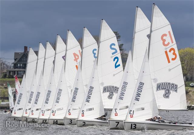 A row of sailboats with 'BROWN' on the sails race closely on the water, their sails numbered and colorful against a cloudy sky, with houses and trees visible in the background.