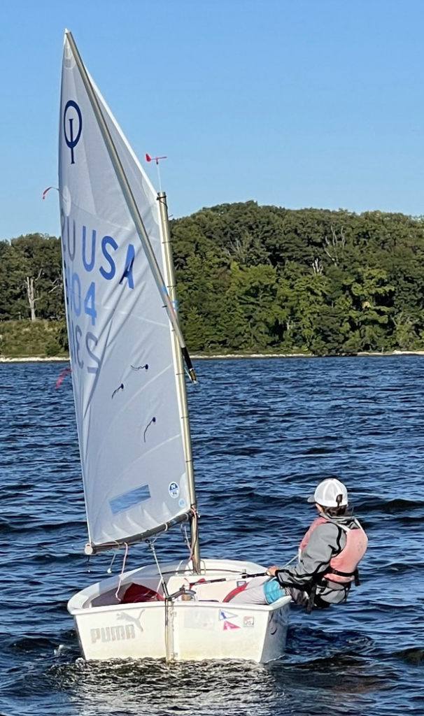 A person wearing a white cap and life jacket sails a small white boat with a tall sail marked "USA" on a lake, with trees and blue sky in the background.