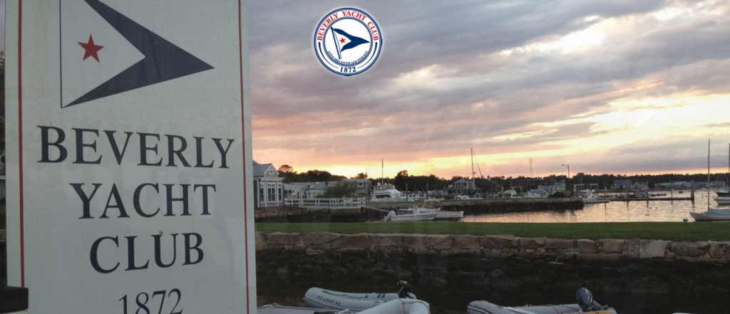 A sign for Beverly Yacht Club, established in 1872, stands by a harbor with boats, a stone wall, and buildings, beneath a cloudy sunset sky. The club's round logo appears above the scene.