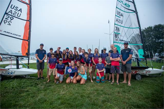 A group of youth and adults pose together in front of sailboats on a grassy area, all smiling at the camera. Some wear life jackets and sporty clothes. The scene looks like a sailing event or camp.