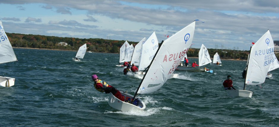 Children sail small dinghies with white sails on a choppy lake under a cloudy sky. One sailor leans far out of their boat, balancing against the wind, while others race nearby. Trees line the distant shore.