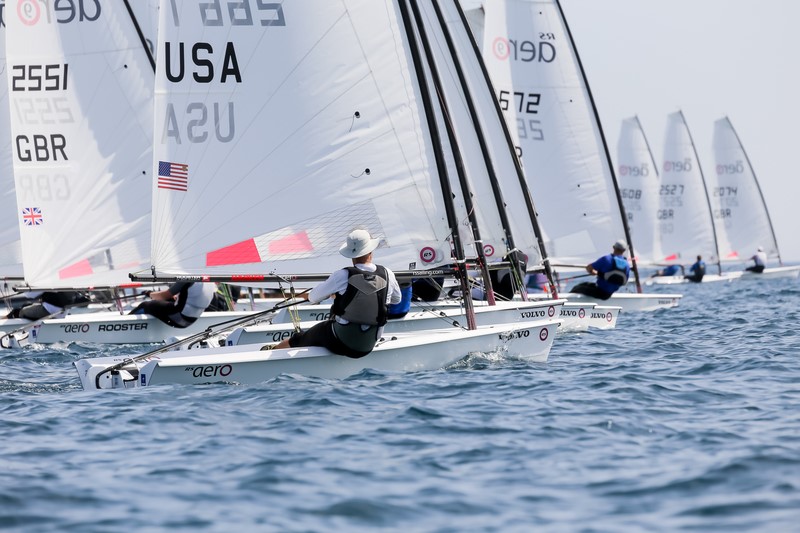 Several small sailboats race side by side on the open water. The sailors are wearing life vests and hats, and the sails display country codes such as USA and GBR. The sky is clear and the water is calm.