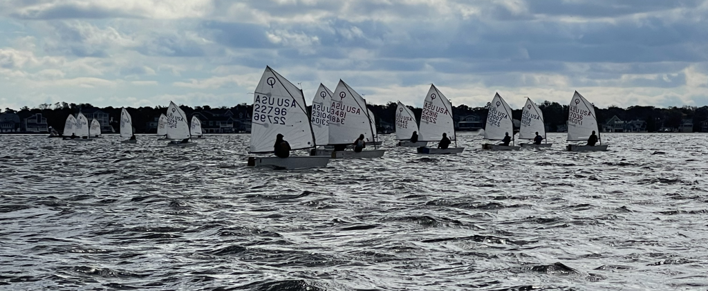 A group of small sailboats with white sails and visible identification numbers glides across choppy water under a cloudy sky, with houses and trees in the background.