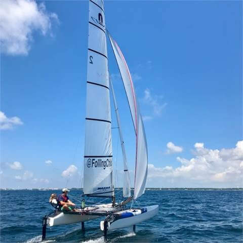 A person is sailing a small catamaran with white sails on blue water under a bright, partly cloudy sky. The boat has '@FoilingClub' written on the sail.