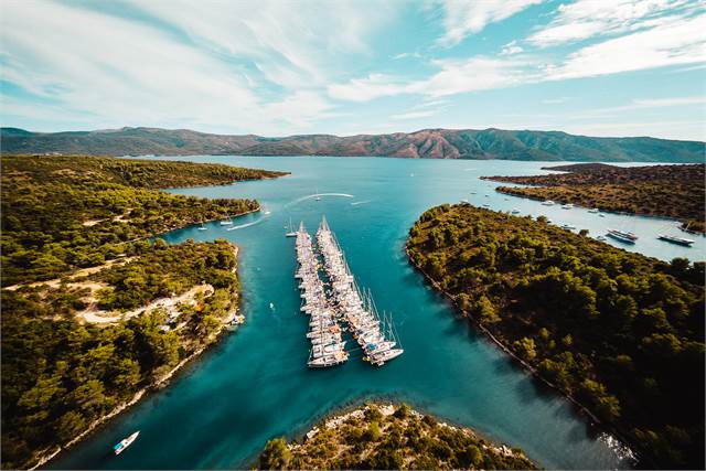 Aerial view of a narrow bay lined with green hills on both sides, filled with numerous sailboats anchored in neat rows, surrounded by calm blue water and distant mountains under a partly cloudy sky.