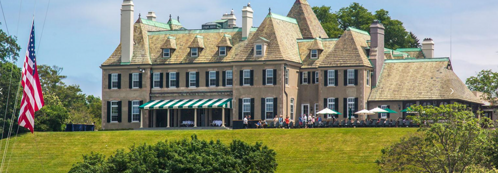 A large, elegant mansion with a tan facade and green roofs sits atop a grassy hill. People are gathered on a terrace with white umbrellas, and an American flag flies on the lawn in front.