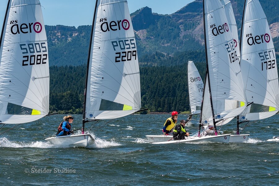 Several people sail small RS Aero racing sailboats on choppy water with forested hills and mountains in the background. The boats' sails display numbers and logos. The sky is clear and sunny.