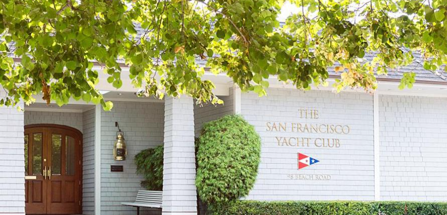 The entrance to The San Francisco Yacht Club, featuring a wooden door, a white bench, manicured greenery, and the club's name and address on a gray shingle wall under leafy green tree branches.