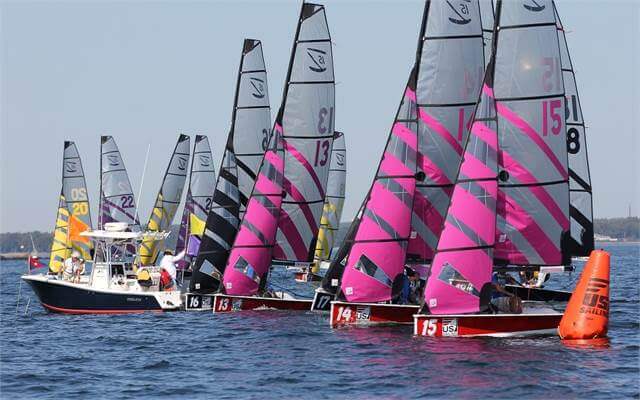 A group of colorful sailboats with pink and yellow striped sails line up at the start of a race on a calm lake, next to a white motorboat and an orange buoy under a clear blue sky.